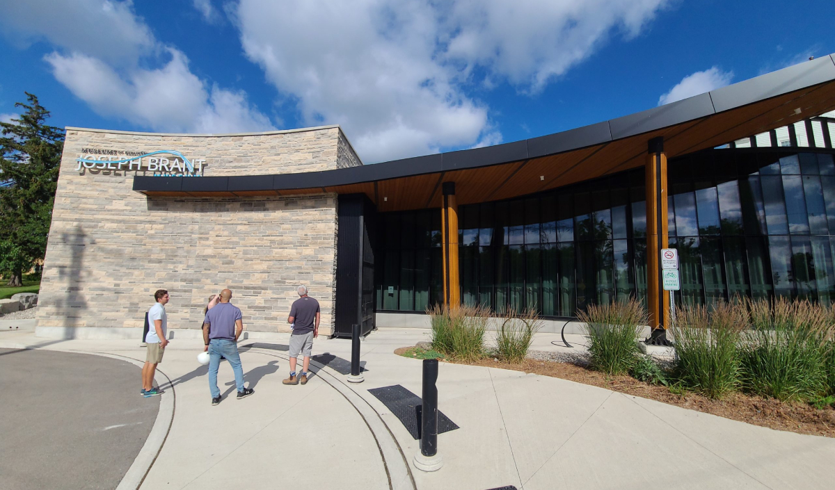 Three people walk toward the entrance of the Joseph Brant Museum, a striking modern stone and glass building under a blue sky with scattered clouds, highlighting the landmark’s inviting presence.