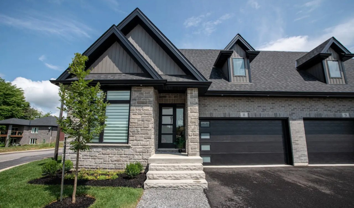 A modern semi-detached house with stone and brick exterior, black roof, large windows, two garages, a small front yard, and a young tree by the entrance.