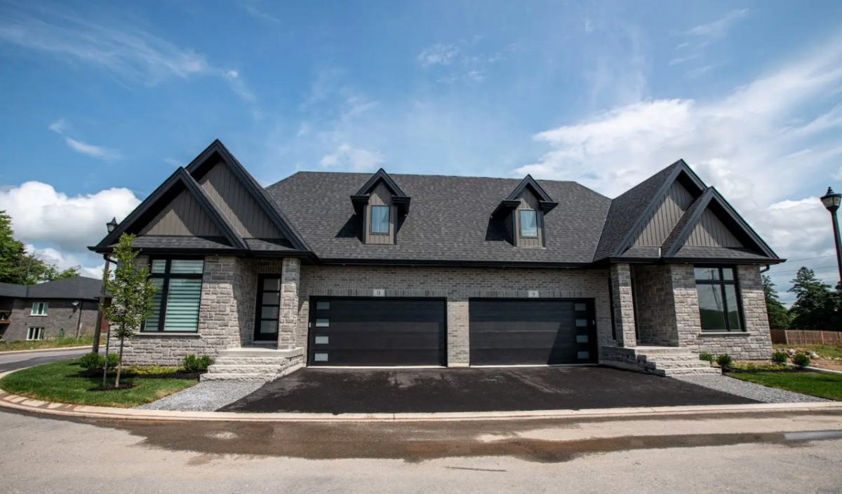 A duplex house with two garages, stone and dark siding exterior, black roof, dormer windows, and a paved driveway under a partly cloudy sky.