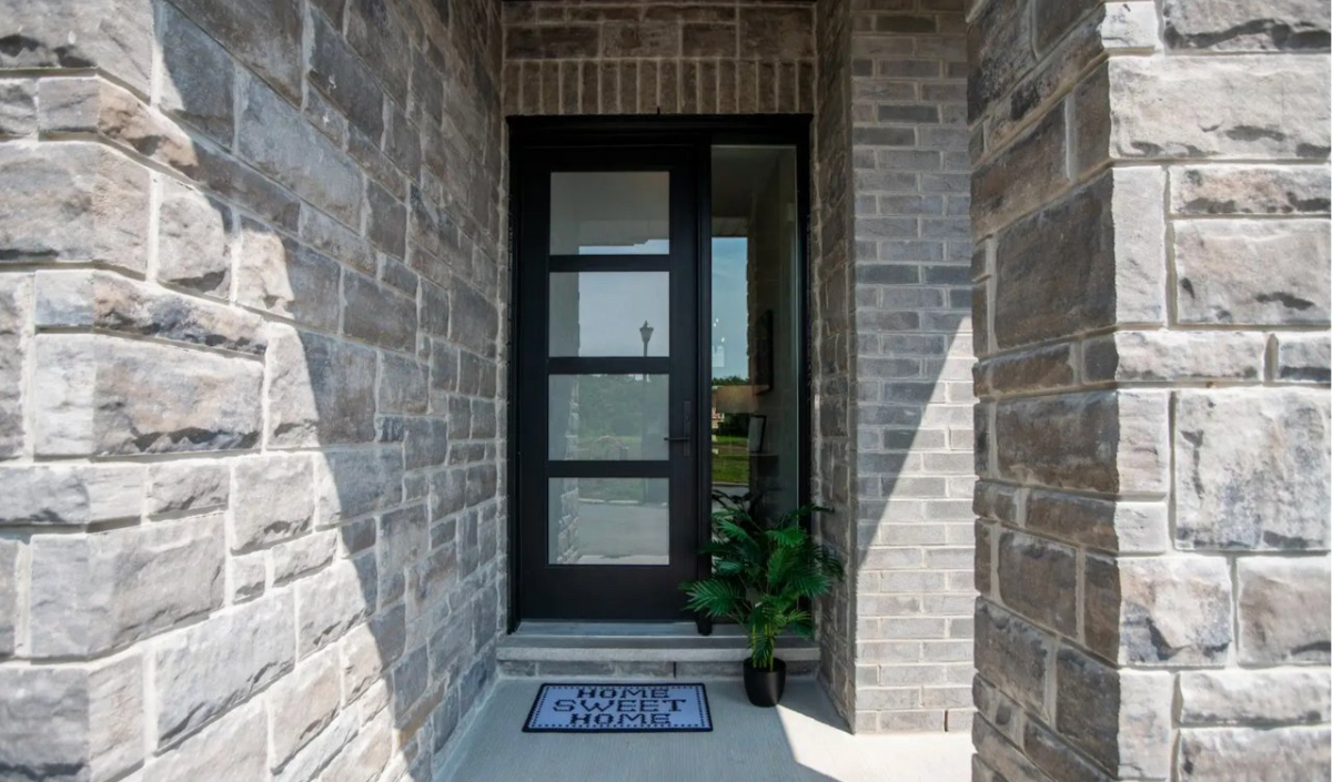 Modern front door with black trim, surrounded by gray stone walls, a "Home Sweet Home" doormat, and a potted plant to the right of the entrance.