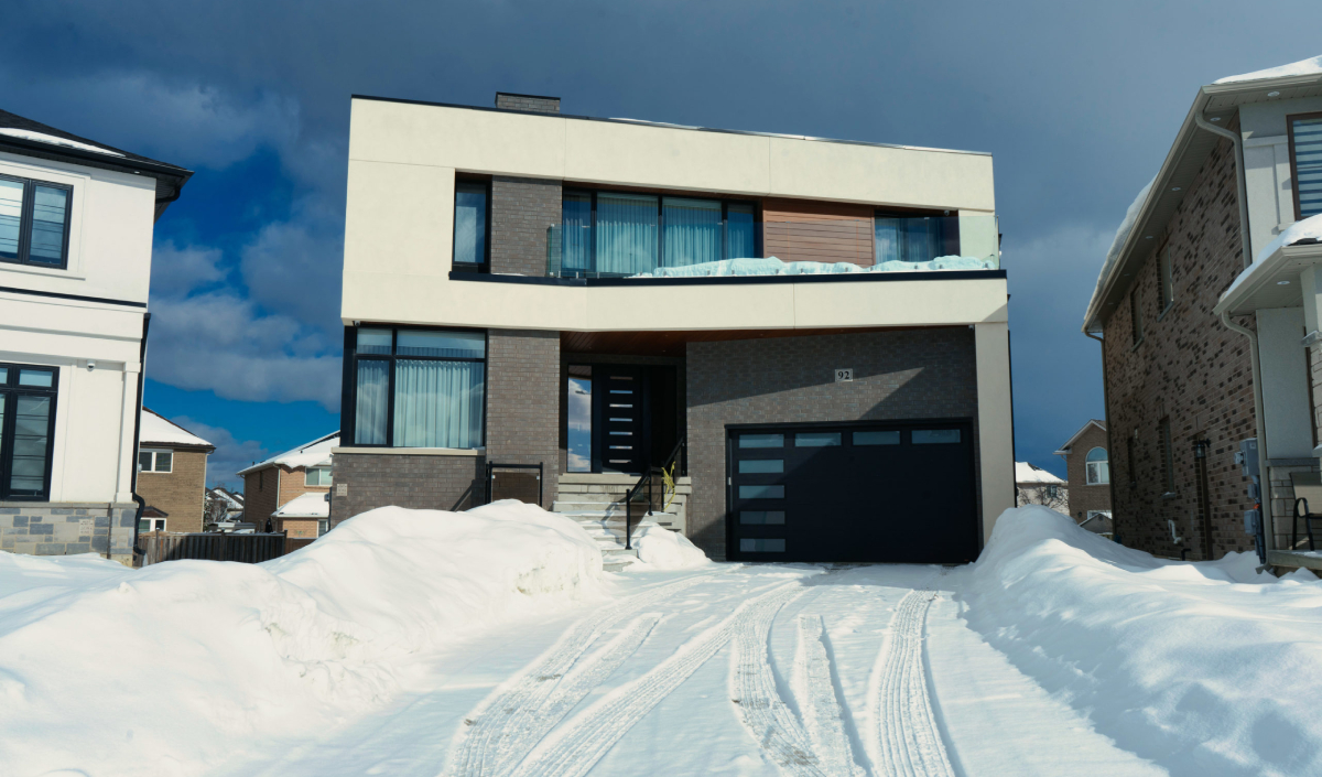 A modern two-story house with large windows and a garage, surrounded by snow on the driveway and steps, under a cloudy sky.