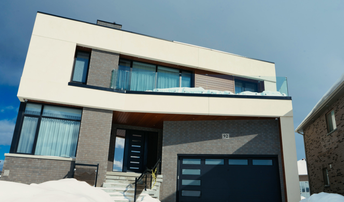 A modern two-story house with large windows, a balcony, a black garage door, and snow on the ground and roof.