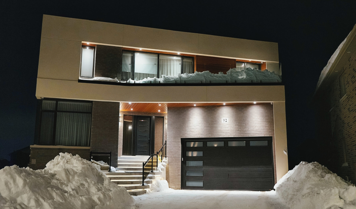 Modern two-story house at night with large windows, an attached garage, snow piled on the ground, and snow accumulated on the balcony railing.