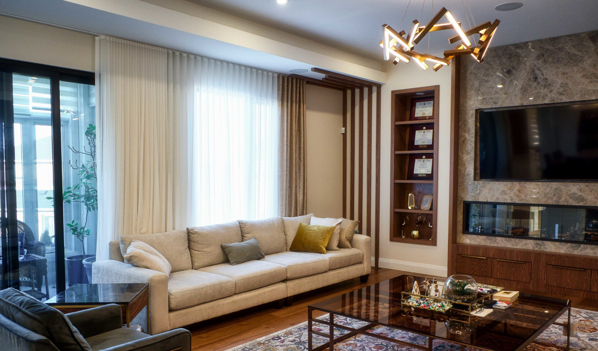 Modern living room with a beige sofa, glass coffee table, wall-mounted TV, geometric light fixture, and built-in shelving displaying awards. Large windows with sheer curtains let in natural light.
