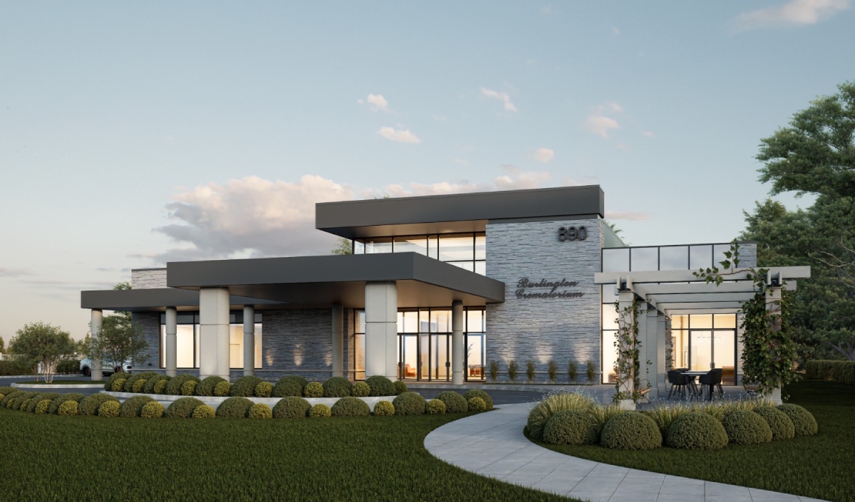 Modern building with large windows, flat roof, stone and metal exterior, surrounded by landscaped bushes, lawn, and a pergola-covered patio area.