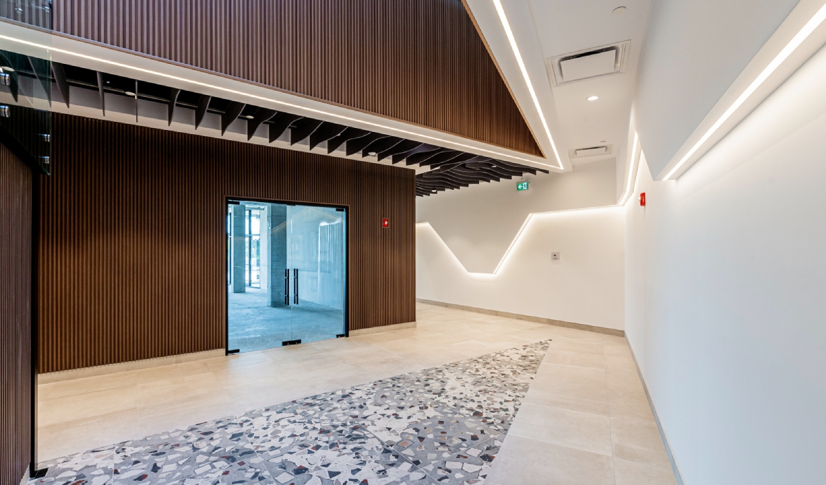 Modern building hallway with geometric ceiling and wall lighting, glass door entrance, and tiled floor featuring a patterned central strip.