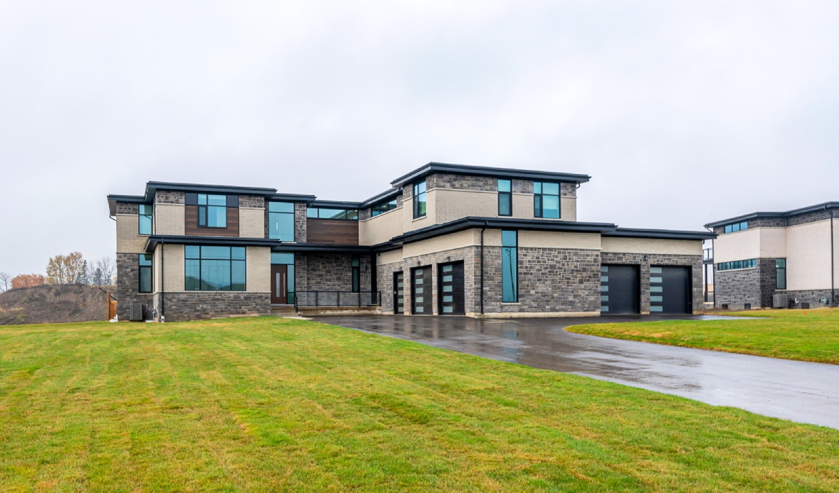 Modern two-story house with large windows, stone and stucco exterior, three-car garage, and a wide driveway on a rainy day.