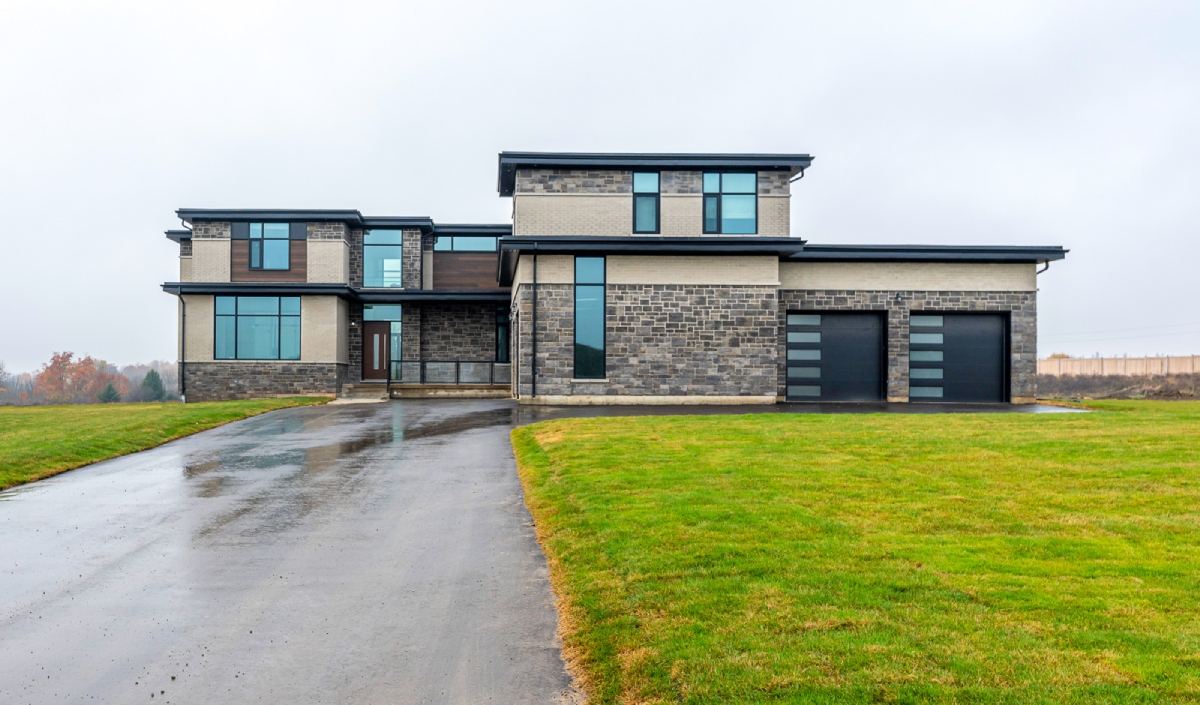 A modern two-story house with large windows, mixed stone and siding exterior, double garage, and a wet driveway on a cloudy day.