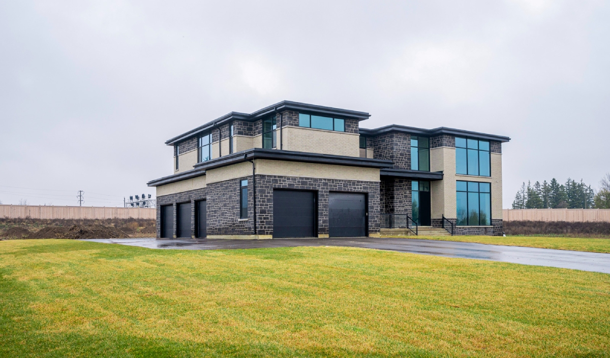 A modern two-story house with large windows, brick and stone exterior, three garage doors, and a wide driveway, situated on a grassy lawn under an overcast sky.