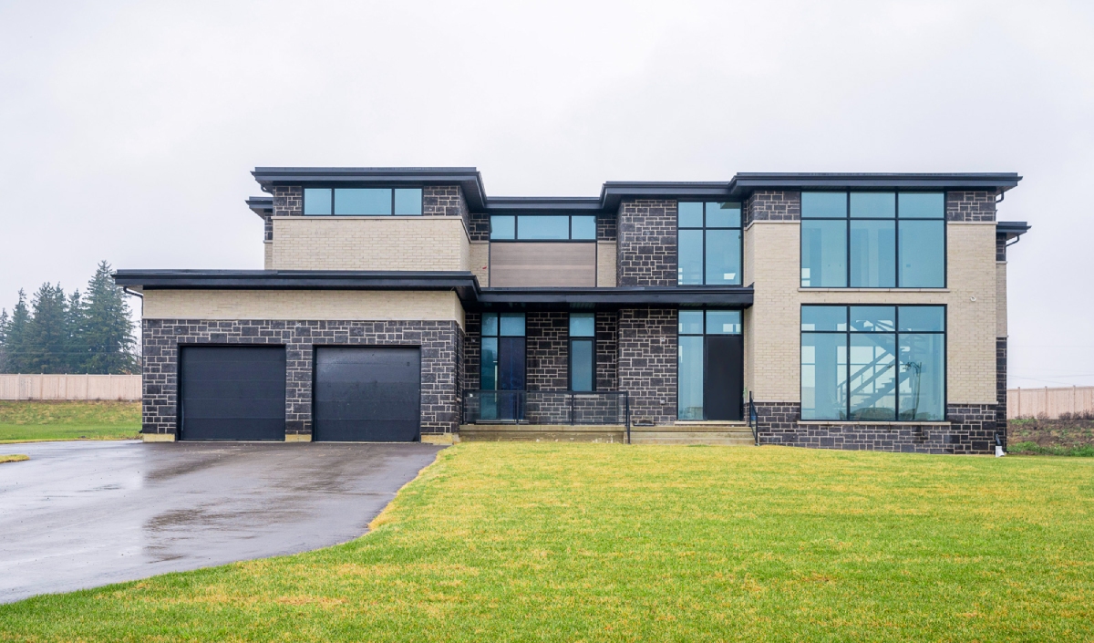 Two-story modern house with large windows, a double garage, and a mix of light brick and dark stone exterior, situated on a grassy lawn with a cloudy sky.