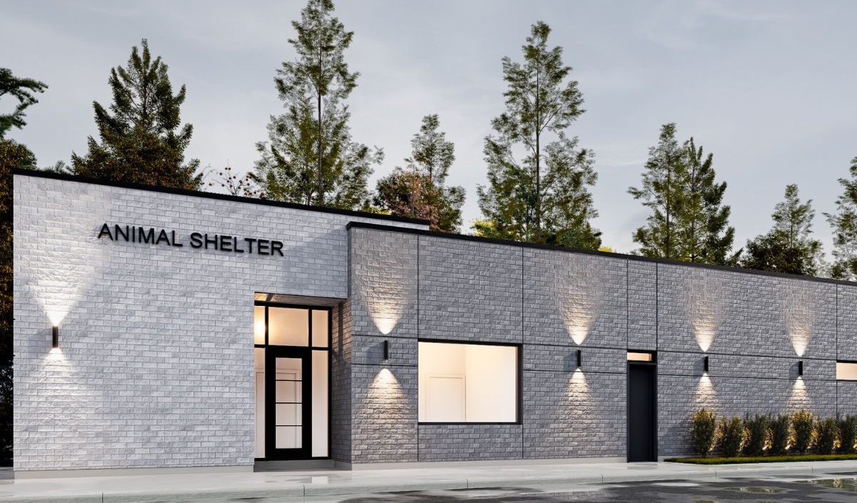 Modern, single-story building with “Animal Shelter” sign, gray brick exterior, large windows, and outdoor lighting, surrounded by trees on a cloudy day.