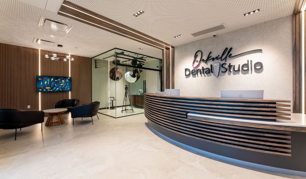 Reception area of a modern dental clinic with a curved desk, black chairs, a small table, wall art, and a glass-walled treatment room in the background.