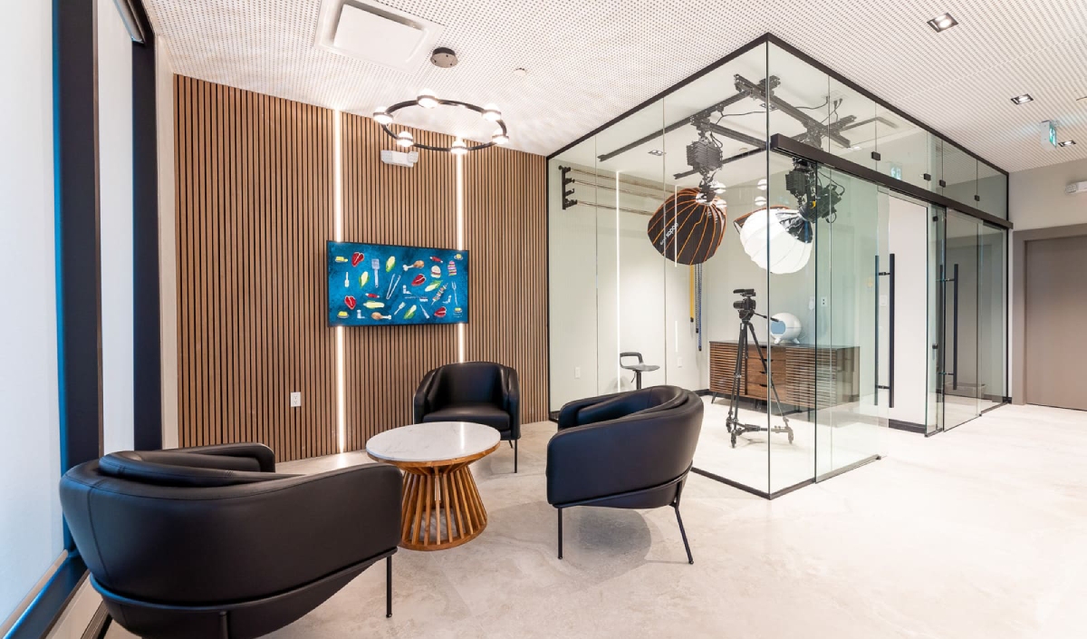 Modern office space with three black chairs around a small round table, a TV on a wood-paneled wall, and a glass-walled studio setup with cameras and lights in the background.