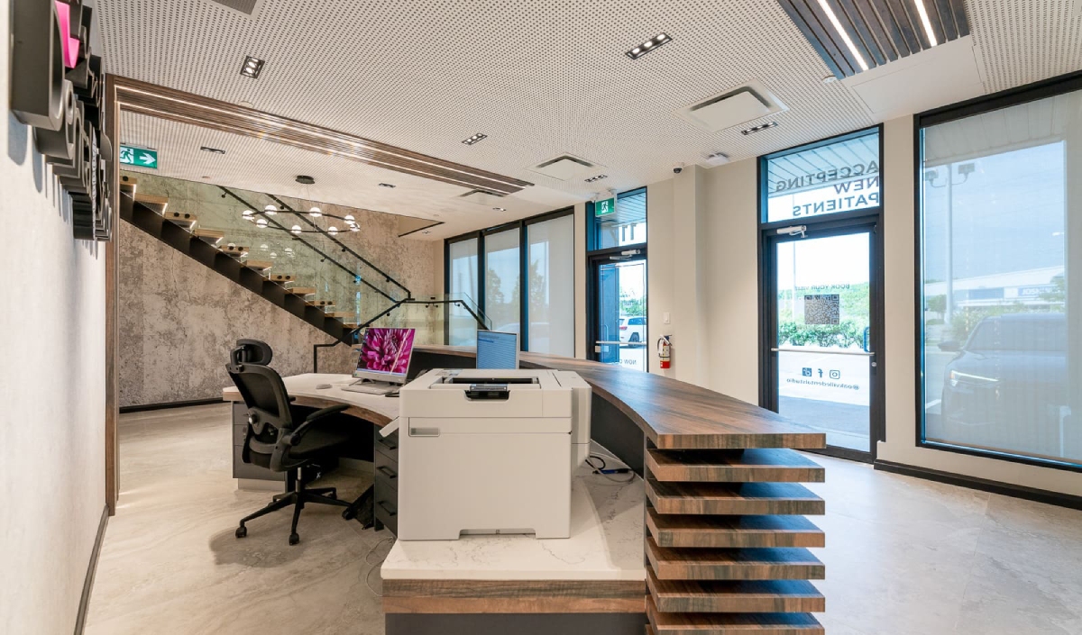 Modern office reception area with a curved wooden desk, computer, printer, large windows, and a staircase with glass railing in the background.