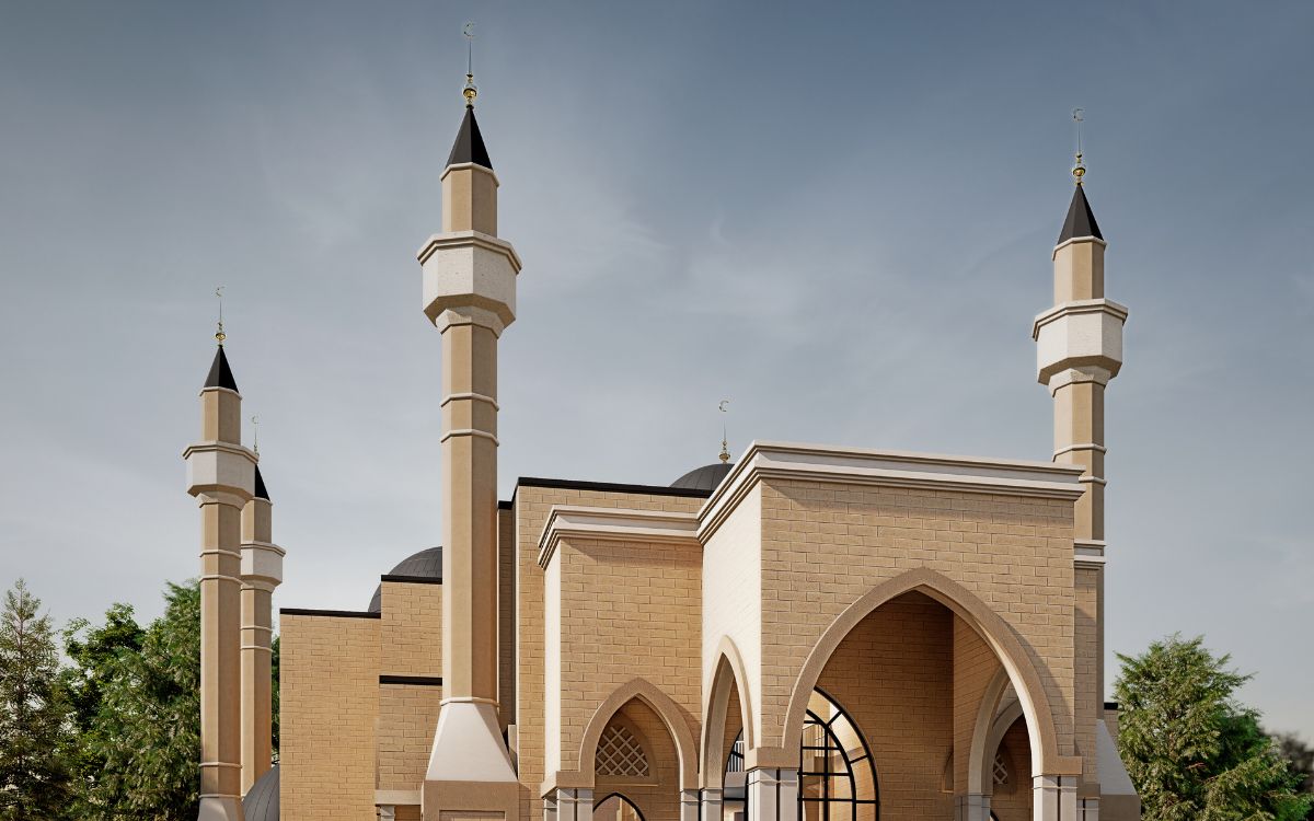 A beige mosque with three tall minarets, arched entrances, and decorative domes, set against a clear sky with some trees in the background.