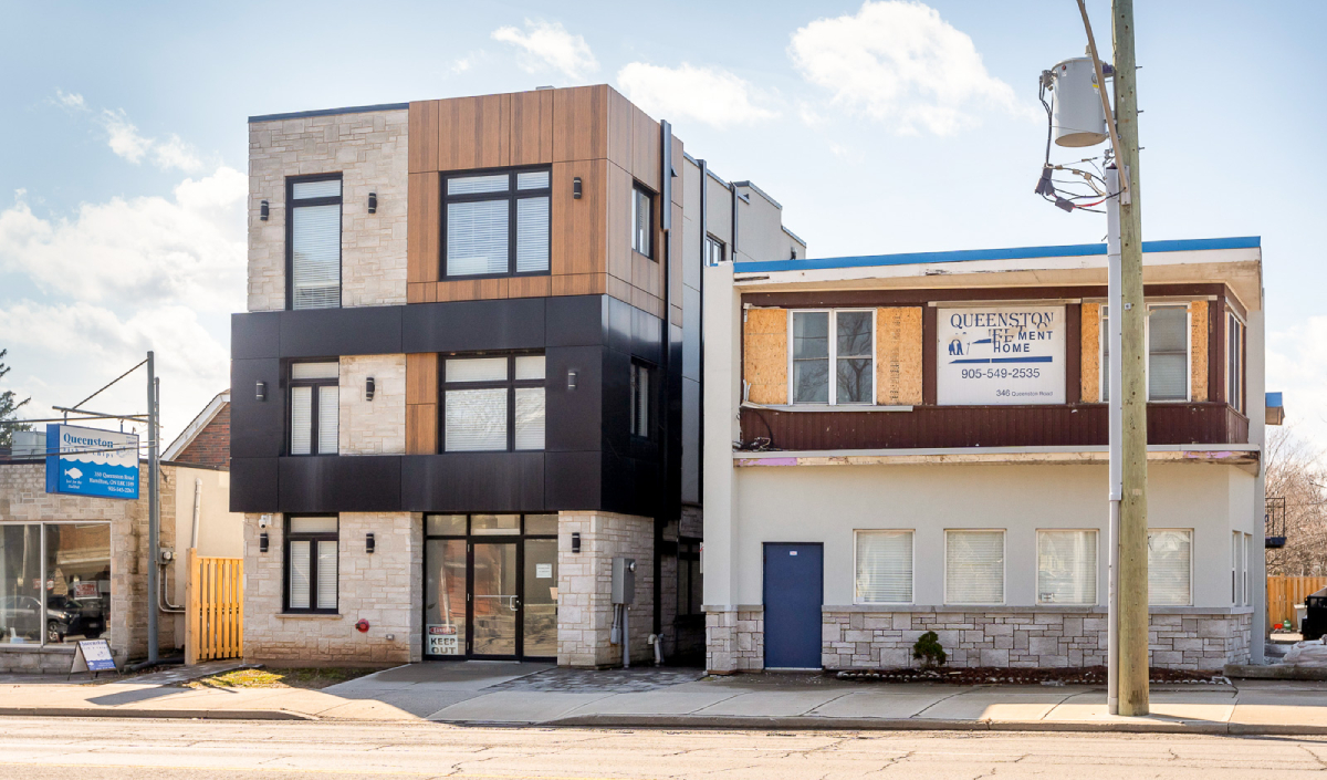 Two adjacent commercial buildings, one modern with wood and black paneling, the other older with a sign reading "Queenston Retirement Home," stand along a main street under a blue sky.