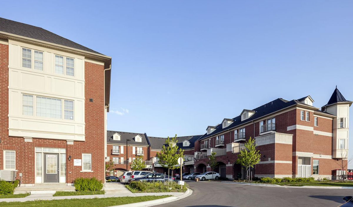 A residential apartment complex with red brick buildings, white trim, multiple windows, and a central driveway lined with parked cars and small trees under a clear blue sky.