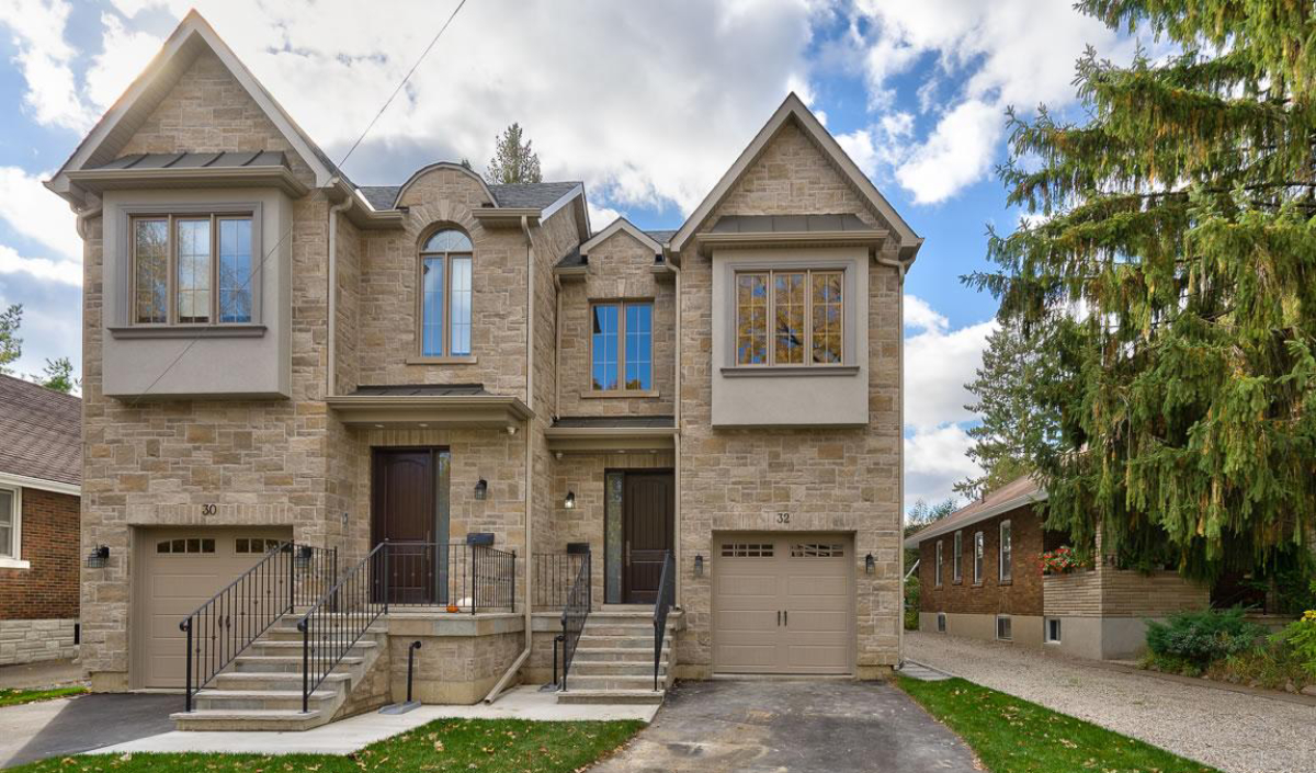 A two-story semi-detached stone house with two front doors, attached garages, and stairs leading to each entrance, set under a partly cloudy sky.