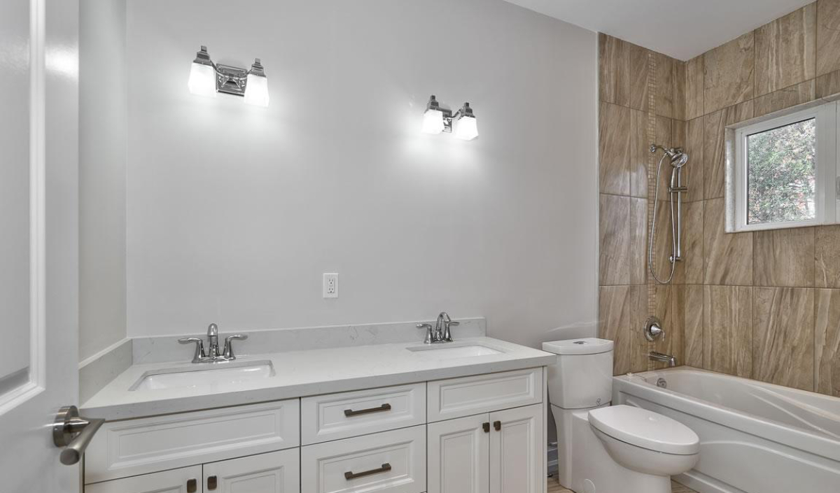 Modern bathroom with double sink vanity, two wall-mounted lights, toilet, and bathtub with tan tile surround and a small window.