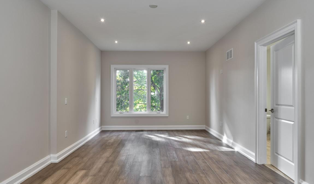 Empty room with light gray walls, wood flooring, recessed ceiling lights, a large window with outdoor greenery visible, and an open white door on the right.