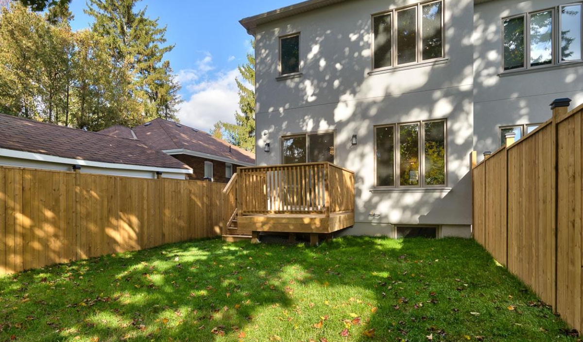 A fenced backyard with green grass, a small wooden deck, and the rear exterior of a two-story house on a sunny day.