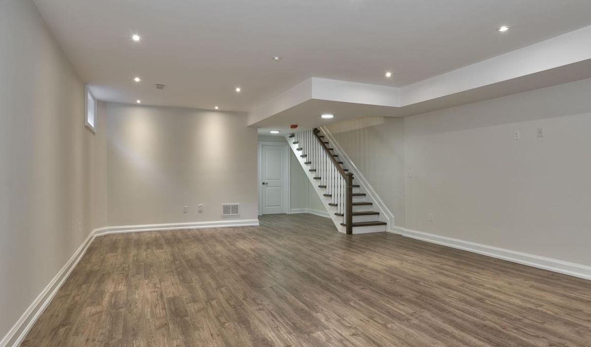 Empty, finished basement with light gray walls, recessed ceiling lights, wood-style flooring, and a staircase leading upstairs in the back right corner.