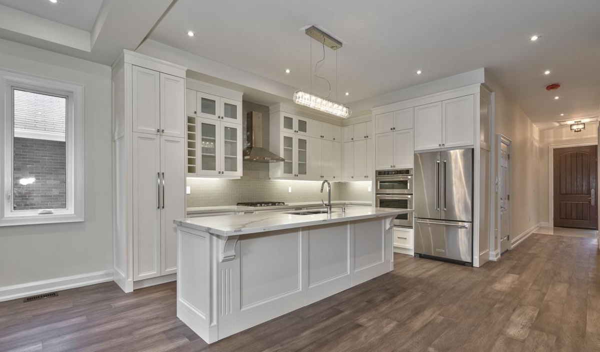 Modern kitchen with white cabinets, a large island with sink, stainless steel appliances, glass-fronted upper cabinets, and wood flooring.