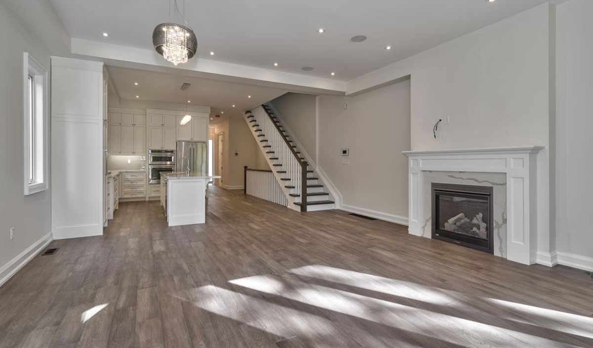 Modern open-plan living area with wood flooring, a white fireplace, an island kitchen, and a staircase leading upstairs. Large windows allow natural light to fill the space.